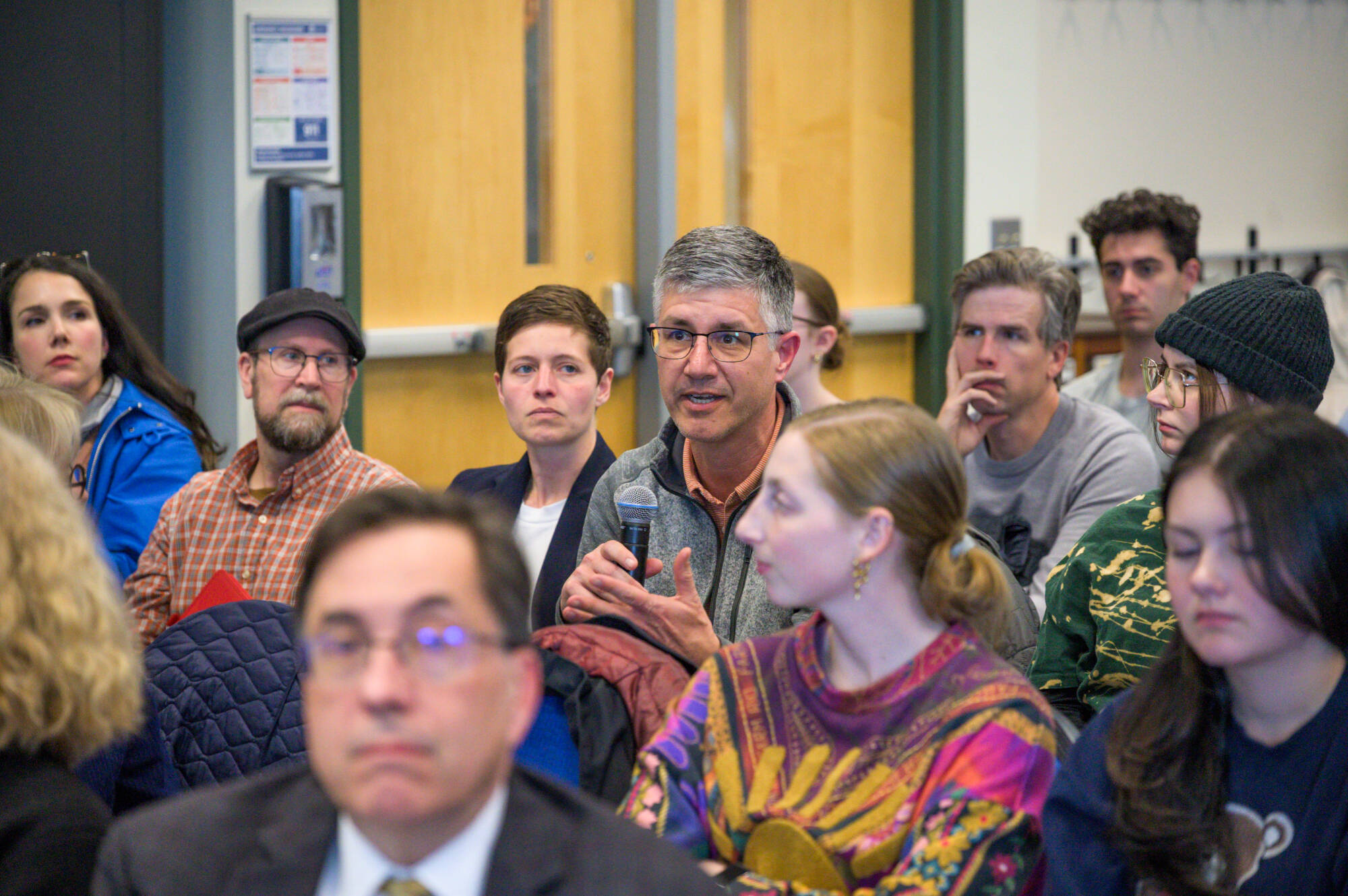 A group of people attentively listen to a speaker holding a microphone during what appears to be a public meeting or seminar.